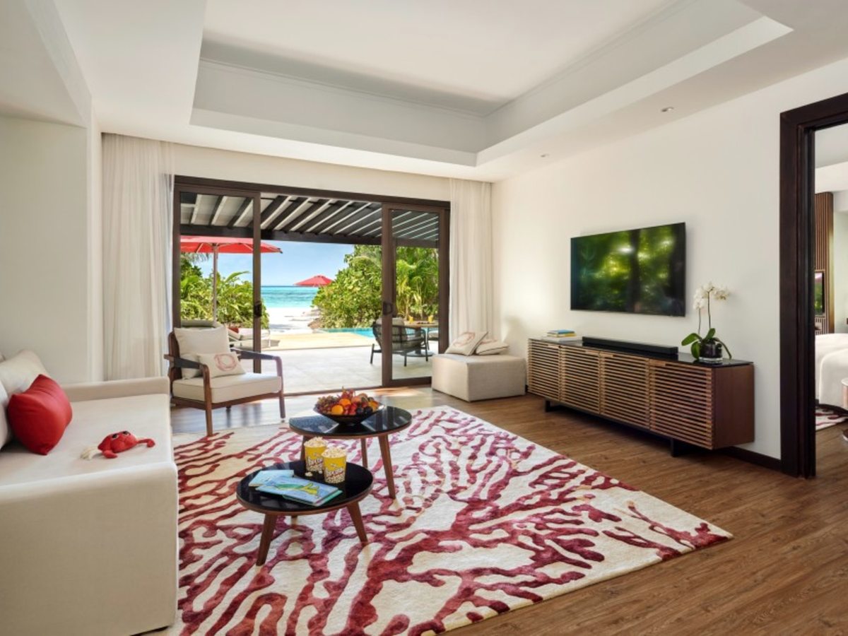 A villa living room with a white sofa, red coral-patterned rug and wall-mounted television, low tables holding popcorn and children's books, and sliding doors open to a pool, a red parasol and the sea.