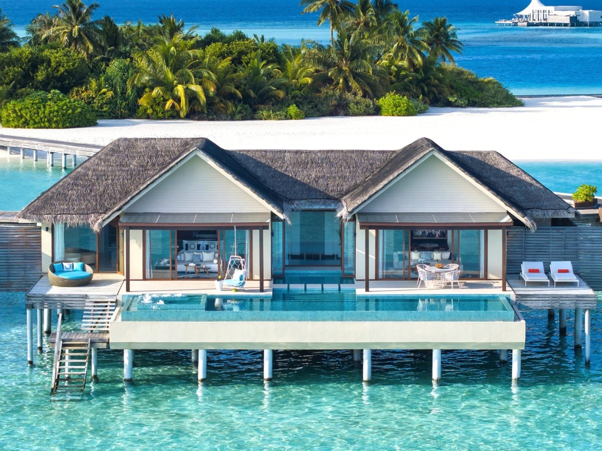 Two linked thatched overwater villa wings on stilts above a clear lagoon, joined by a long swimming pool along the front deck, with a white-sand beach and palm-covered island behind.