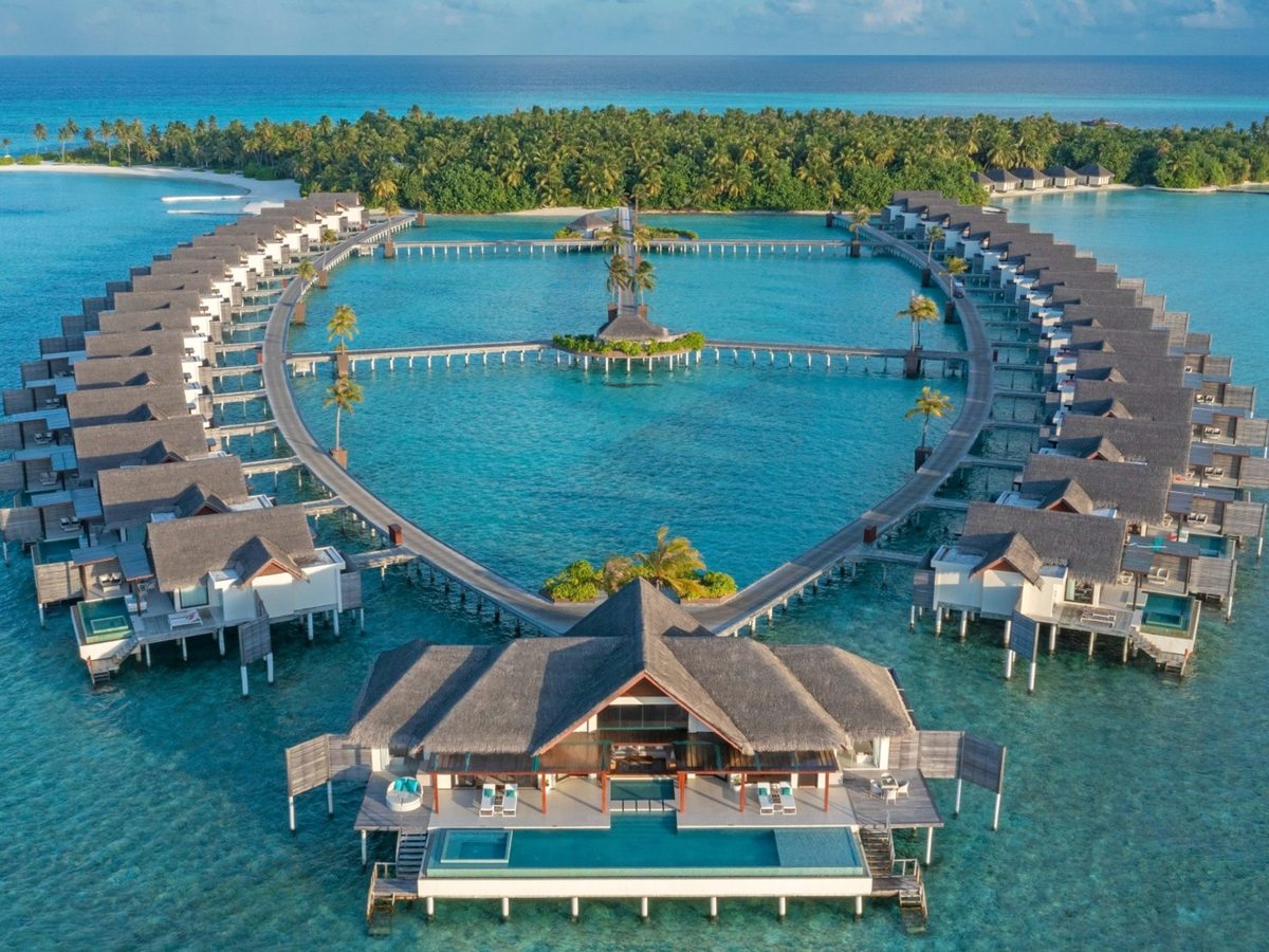 Aerial view of two curving lines of thatched overwater villas on stilts, linked by boardwalks around a turquoise lagoon, with a larger pavilion in the foreground and a palm-covered island behind.