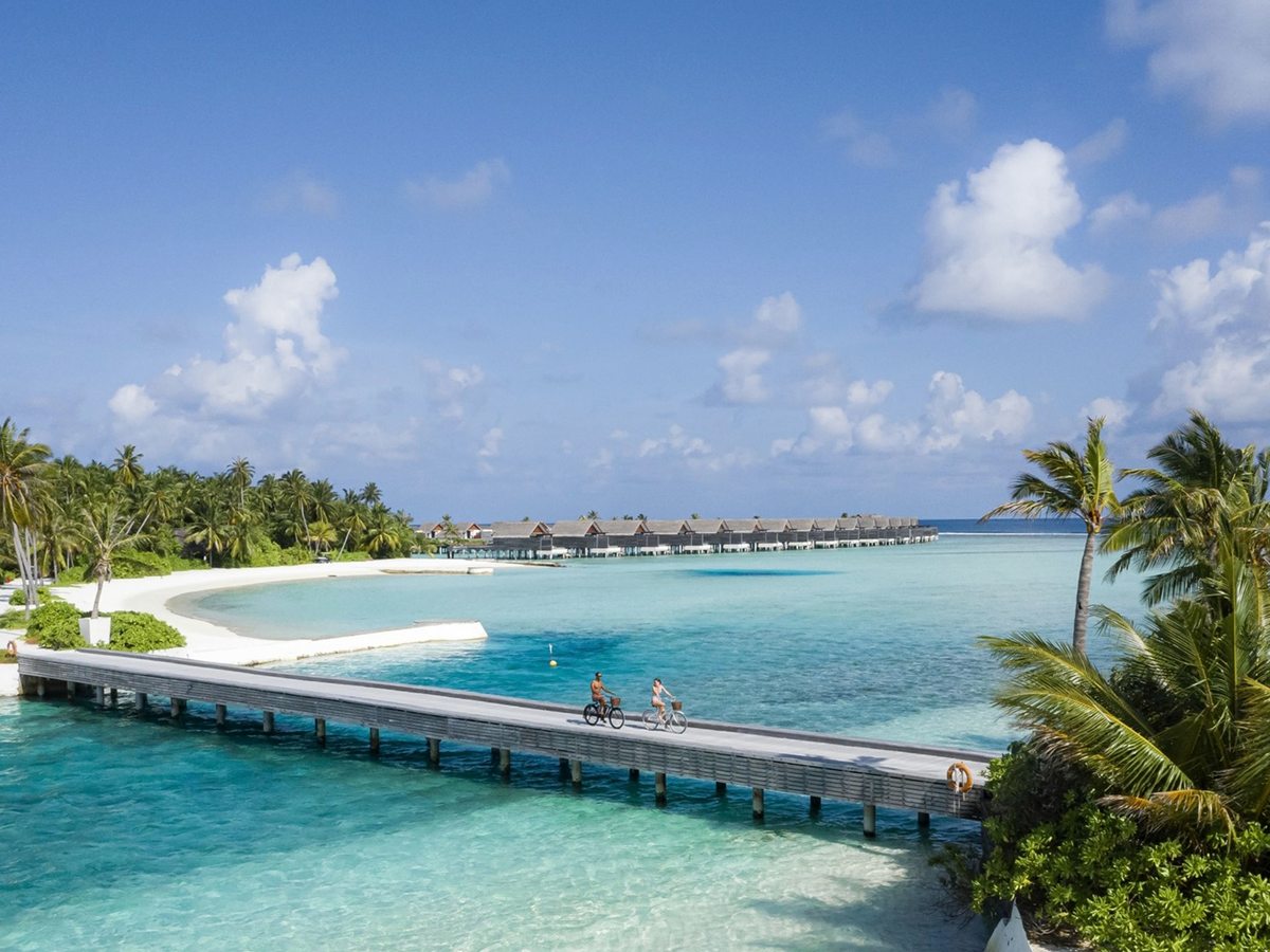 A long timber boardwalk on stilts crossing a shallow turquoise lagoon, with two people cycling along it, a white-sand beach and palms to one side and a line of overwater villas in the distance.