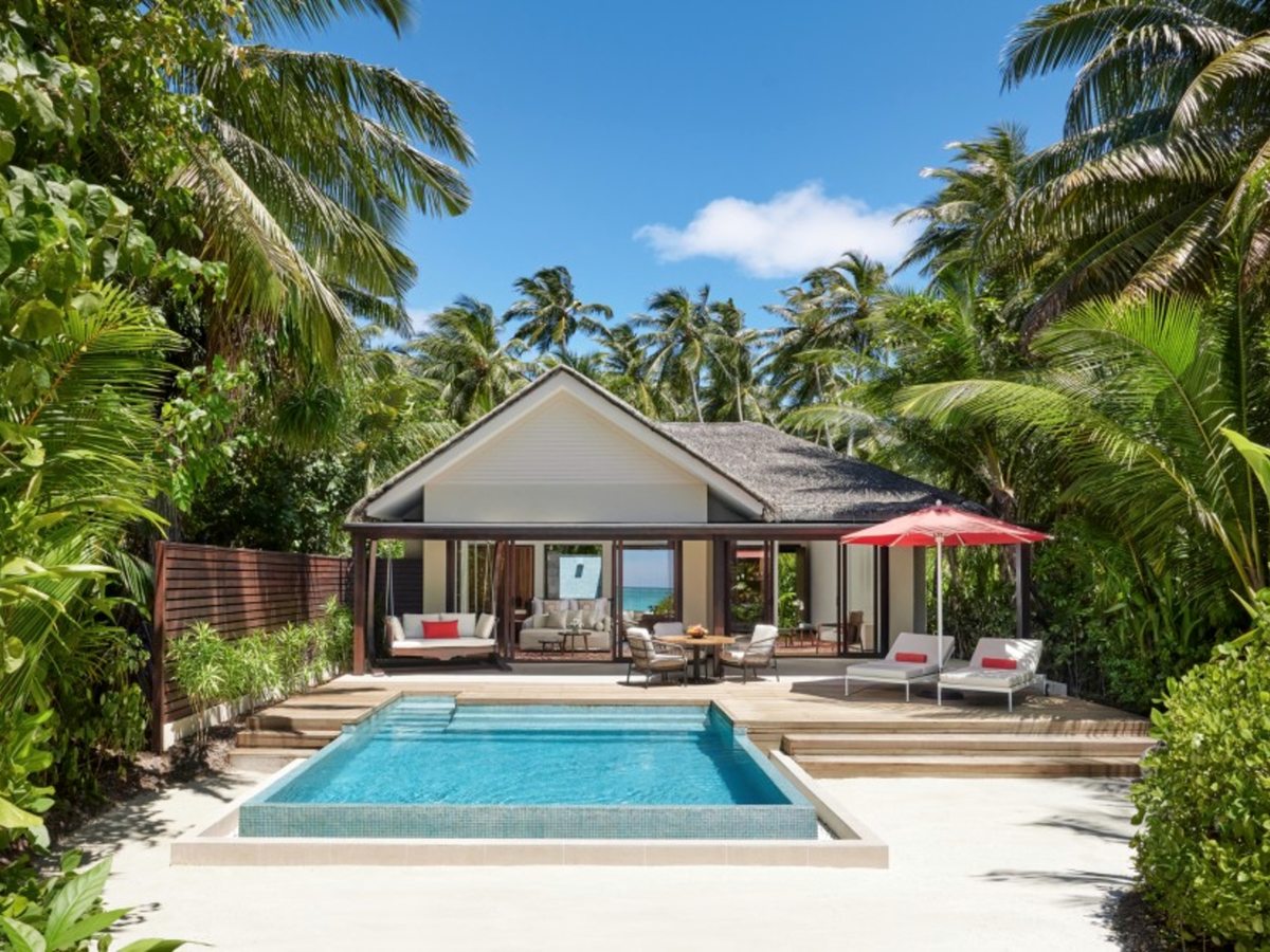 A rectangular plunge pool on white sand in front of a thatched villa with an open living area, a red parasol and two sun loungers, surrounded by palms and greenery.
