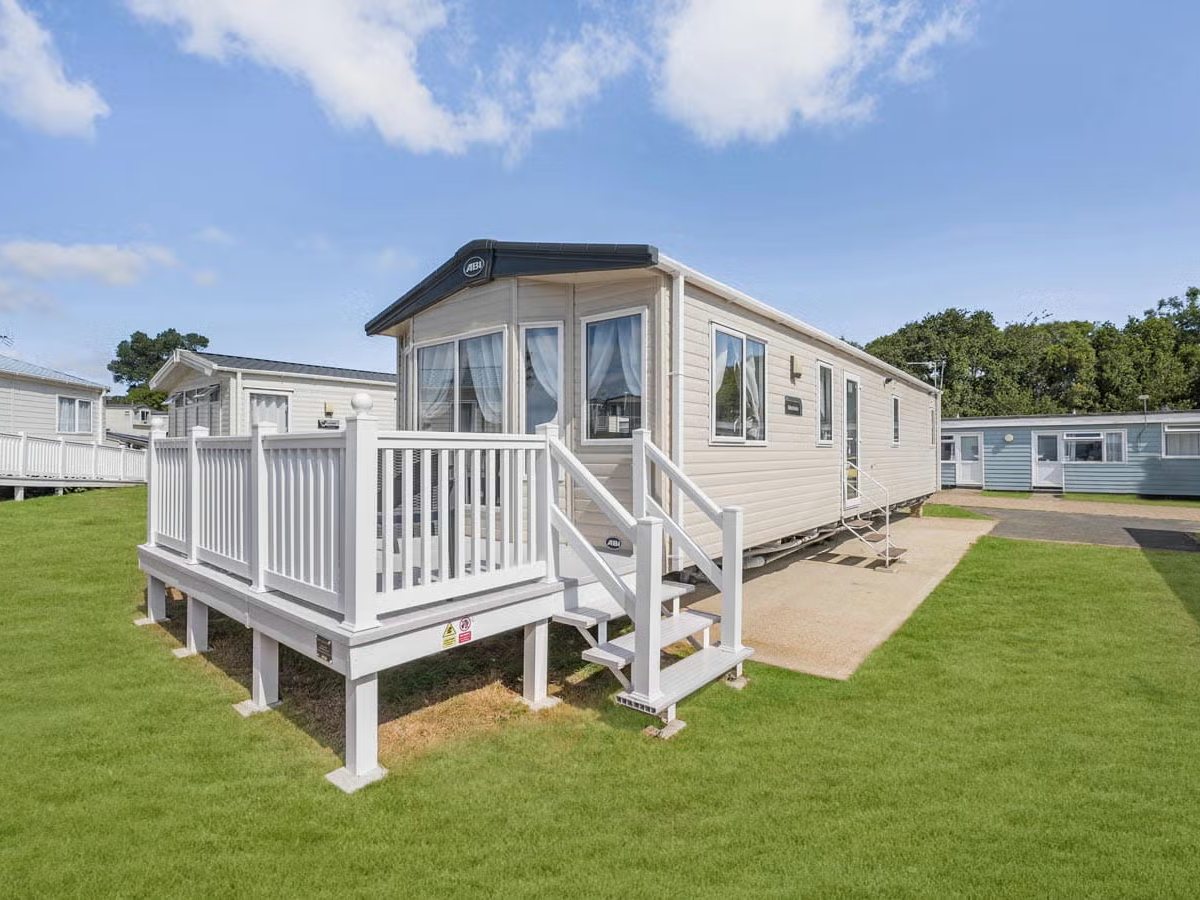 Cream static caravan on a mown grass pitch with a white railed veranda and steps, neighbouring caravans either side under a blue sky.