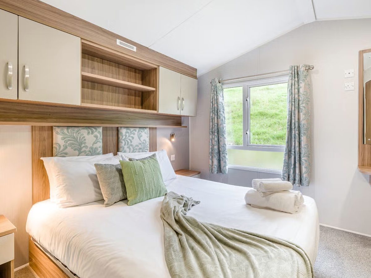 Double bedroom with a white duvet, patterned headboard panels, overhead cupboards, a curtained window and folded towels on the bed.