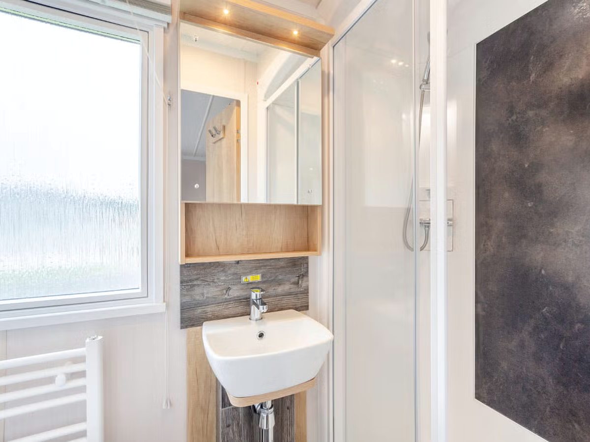 Caravan shower room with a corner shower cubicle, wall-mounted basin, mirrored cabinet, heated towel rail and a frosted window.