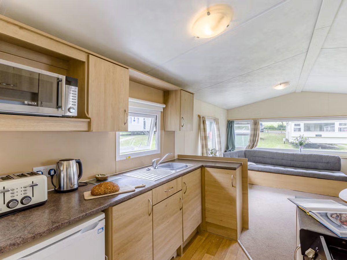 Galley kitchen along one wall of a caravan with sink, worktop, microwave, kettle and toaster, looking through to bench seating and windows.