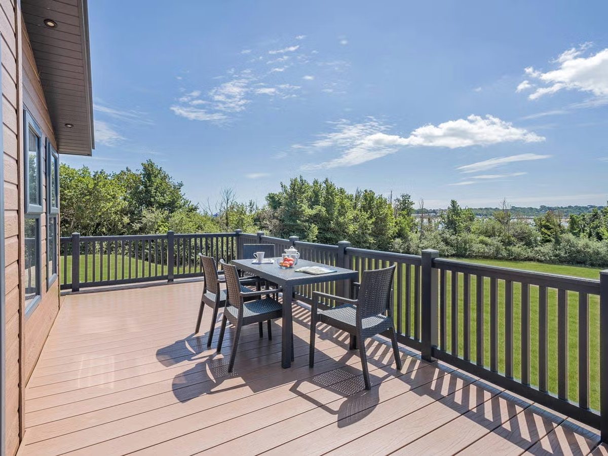 Timber decking with a rattan table and four chairs behind dark railings, lawn and trees beyond and water in the distance.