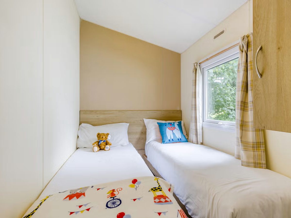 Twin bedroom with two single beds, a teddy on one pillow, a patterned children's throw, a wardrobe and a window onto greenery.