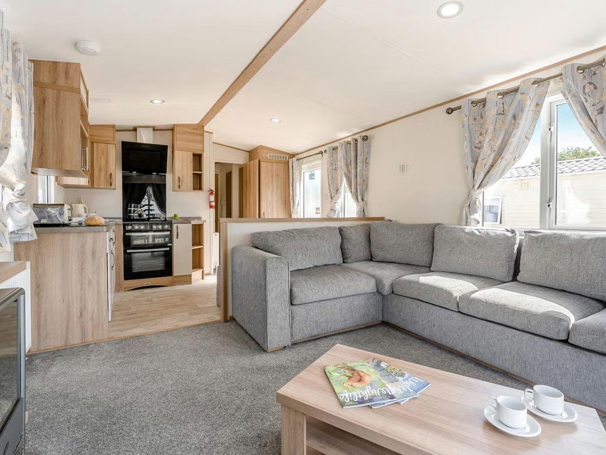 Caravan living area with a large grey corner sofa, wooden coffee table, curtained windows and a fitted kitchen with an oven behind.