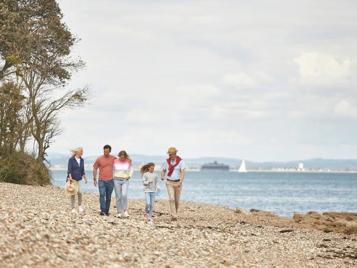 A family walking along the shingle and sand beach below the park, with calm sea, a ferry passing offshore and the mainland coast beyond.