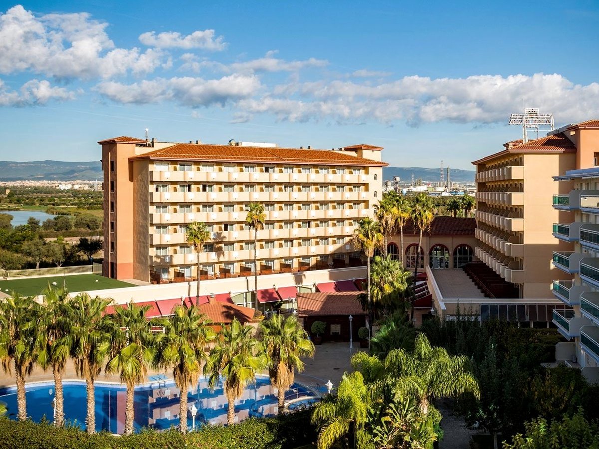 Elevated view of the two apricot bedroom wings meeting at a red-tiled central pavilion, with red awnings over the terrace below, the outdoor pool among palms in front, and trees, an industrial plant and low hills in the distance behind.
