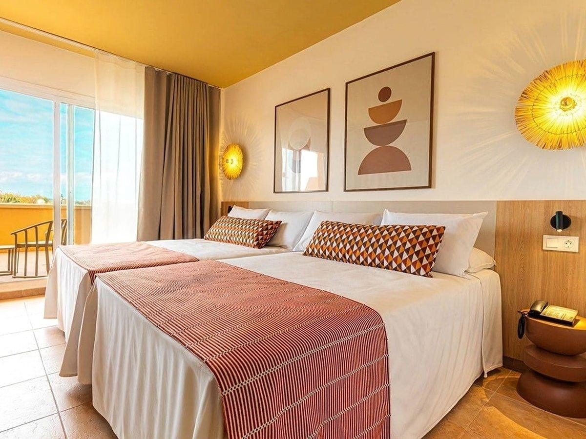 Twin beds pushed together with white bedding, terracotta runners and patterned bolster cushions, framed abstract prints and sunburst wall lights above, and glazed doors open onto a tiled balcony.