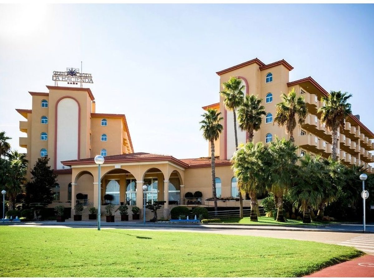 Apricot-rendered hotel with red-tiled roofs and an arched entrance portico, palm trees along the front and a wide green lawn in the foreground under a clear sky.