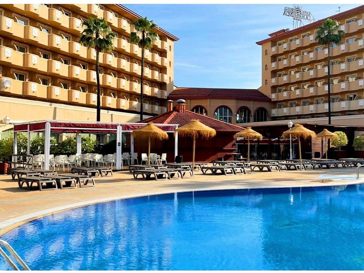 Large outdoor pool with a cream tiled surround, rows of grey sun loungers under straw parasols, and the two apricot hotel wings and red-awninged terrace behind.
