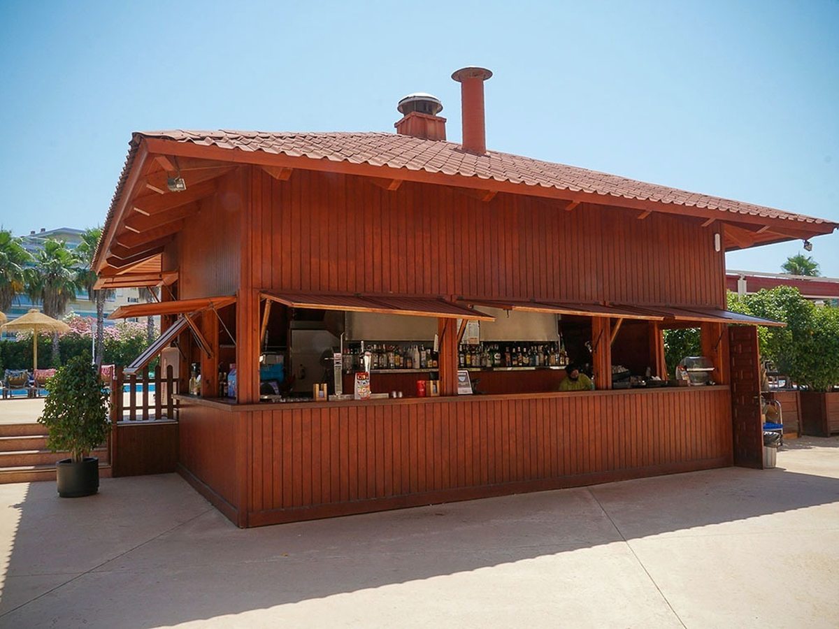 Freestanding timber pool bar with a tiled pitched roof and shutters propped open, spirit bottles lined along the back counter, with sun loungers and the pool behind it.