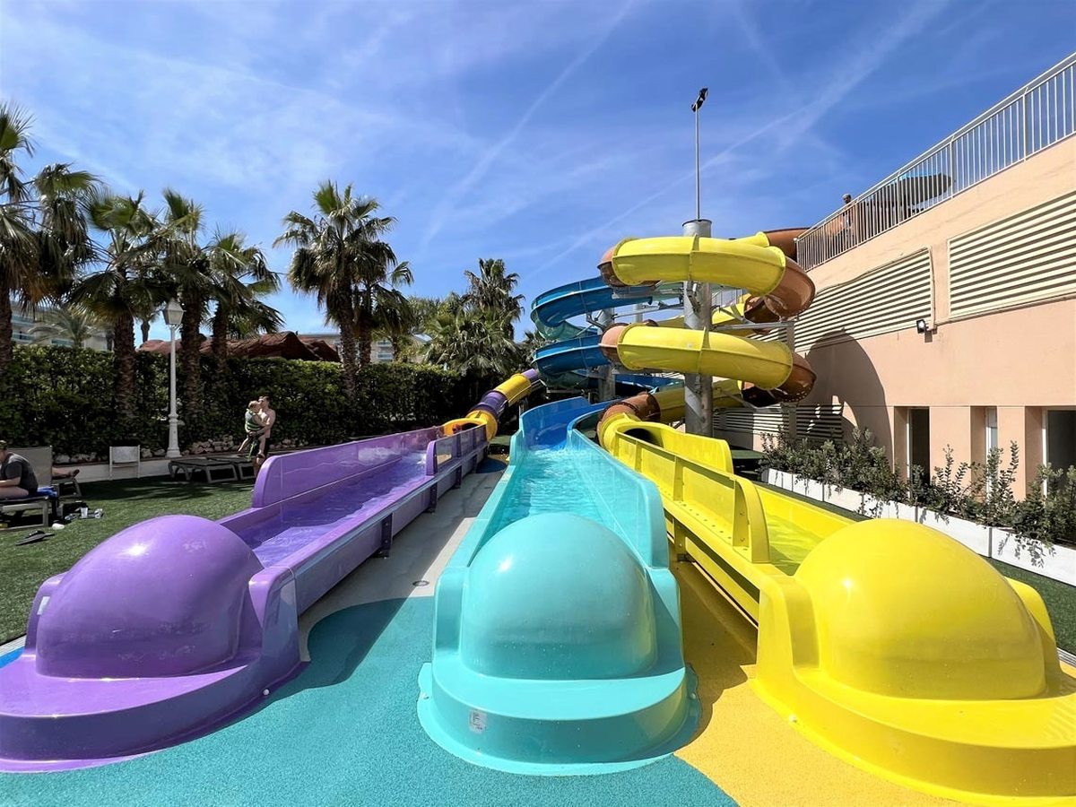 Three wide racer slides in purple, turquoise and yellow running down to their run-out lanes, with two spiral tube slides winding above and palm trees to one side.