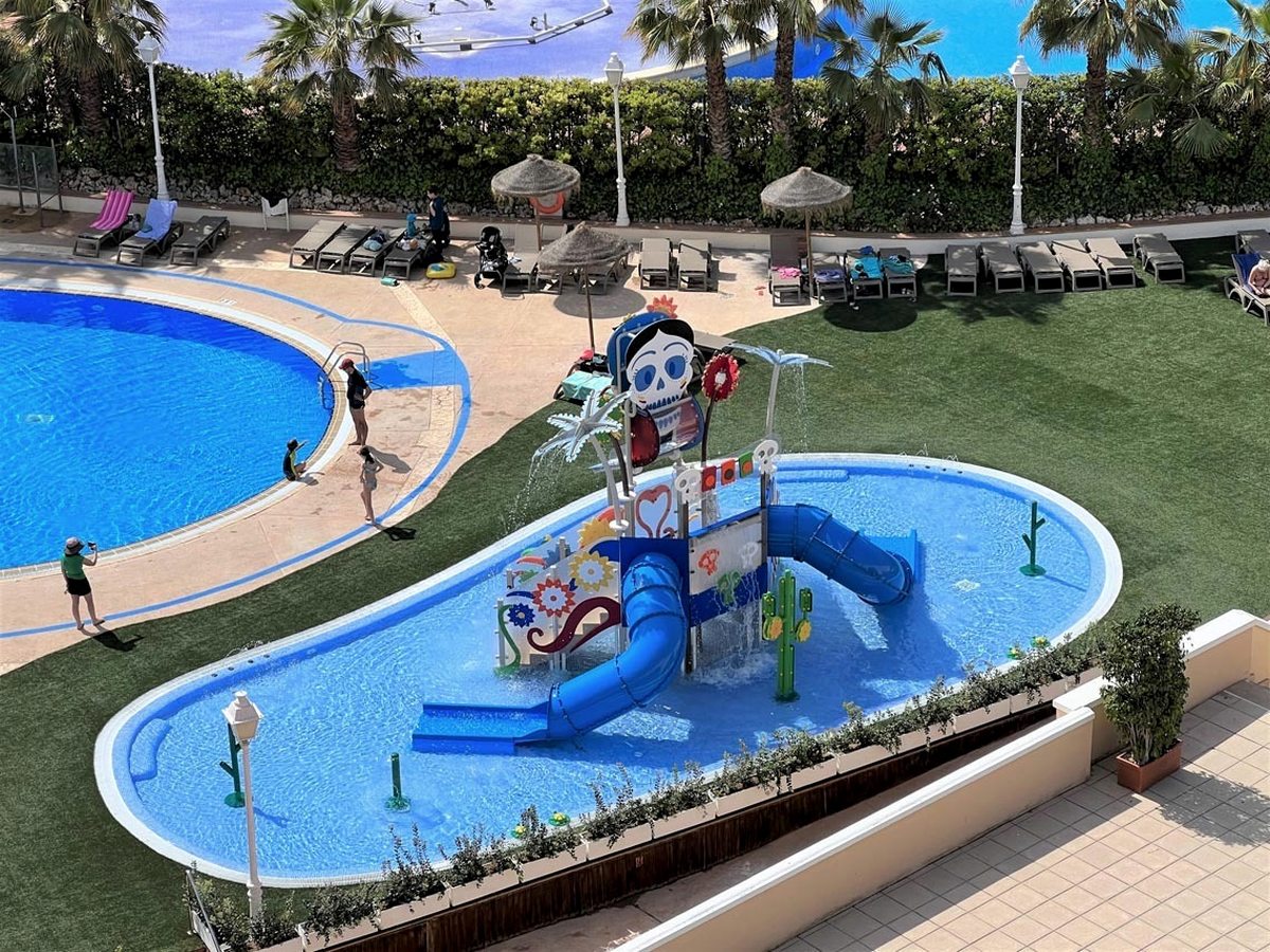 Elevated view of a round shallow children's pool beside an oval splash pool holding a play structure with a blue tube slide and green cactus sprays, set in artificial grass with loungers and parasols beyond.