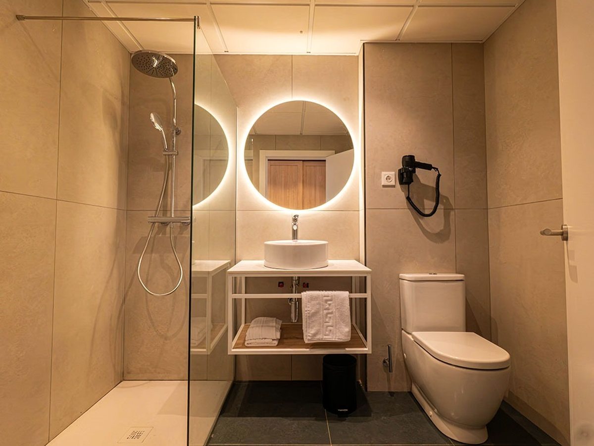 Beige-tiled bathroom with a glass-screened walk-in shower, a round backlit mirror over a white basin on an open timber shelf unit, folded towels beneath and a wall-mounted hairdryer.