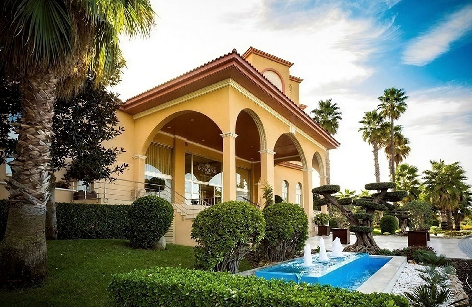 The arched entrance pavilion at Ohtels La Hacienda, in apricot render with a red-tiled roof, with clipped topiary and a low fountain in the garden in front of it.