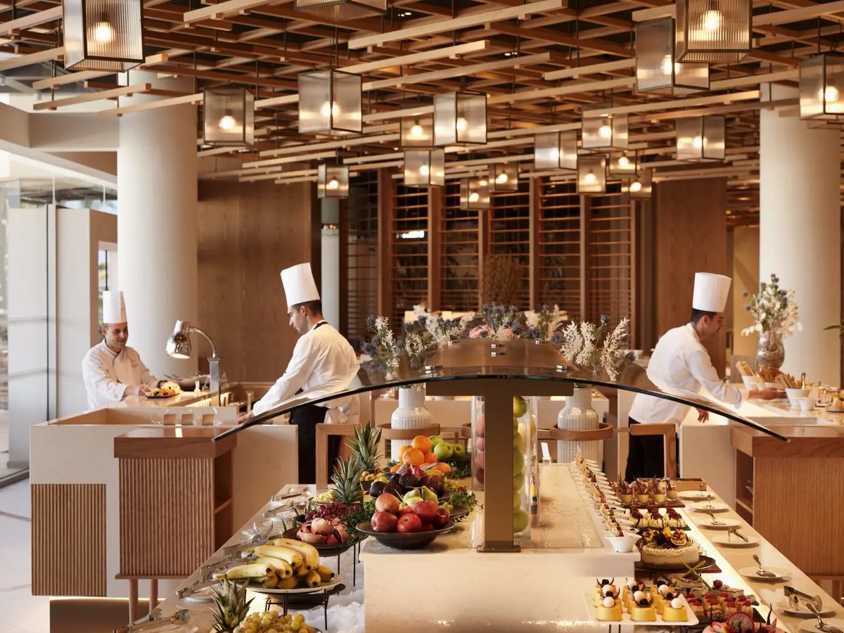 Curved buffet island stacked with fruit, pastries and desserts, three chefs in tall white hats working behind it, under a timber-slatted ceiling of pendant lights.