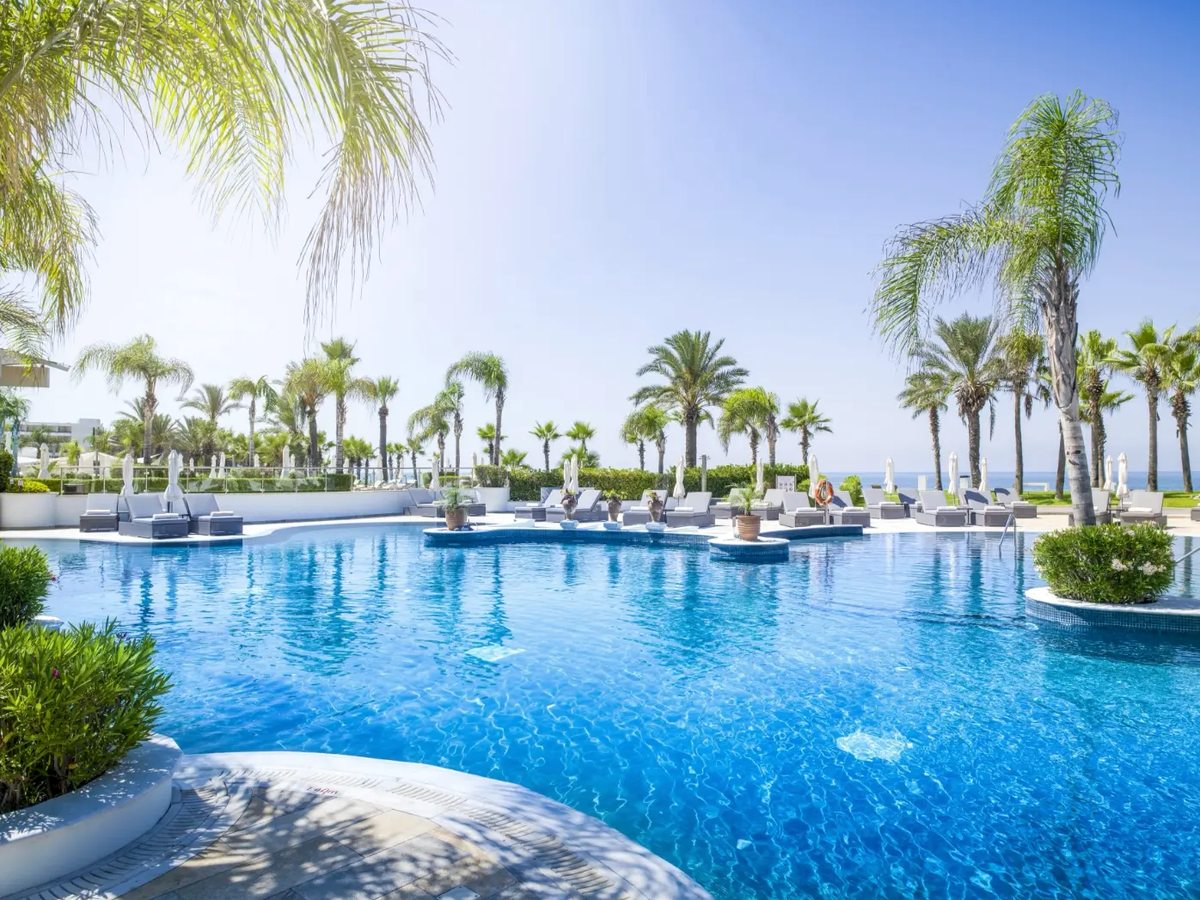 Curved lagoon pool with a planted island, day beds and sun loungers along the far edge, tall palms and the sea beyond.