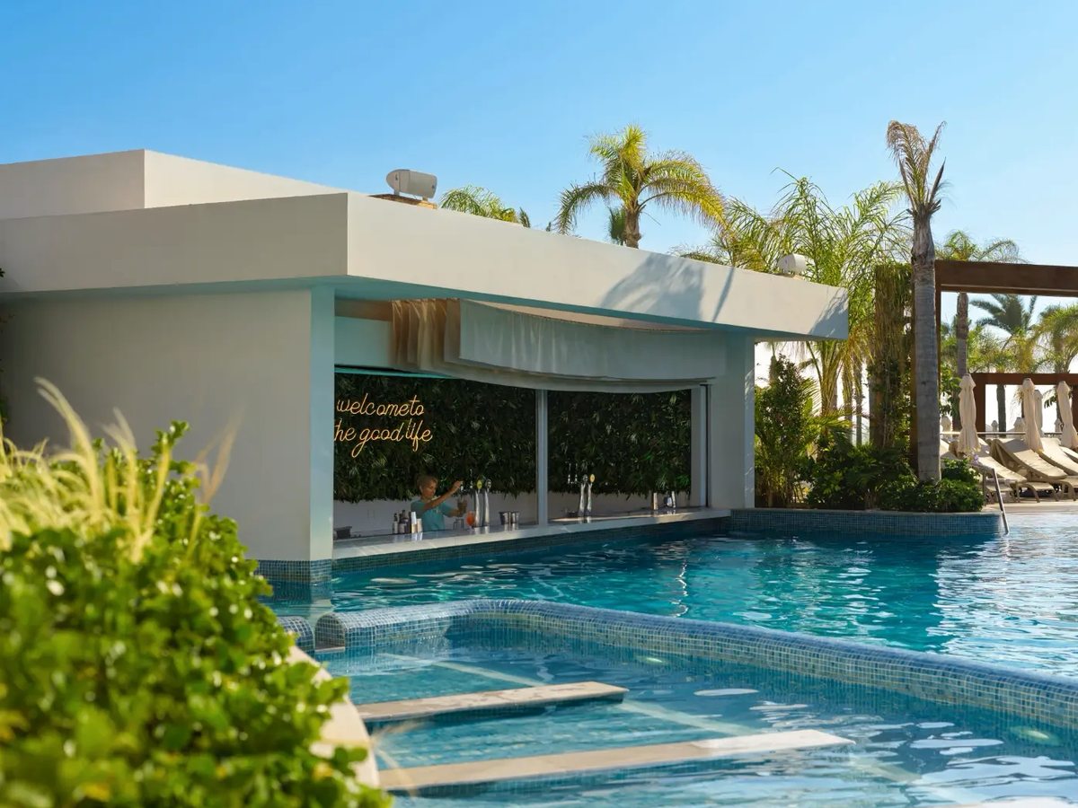 White single-storey poolside bar with a planted wall and a painted sign, the blue pool running up to its counter and palms behind.