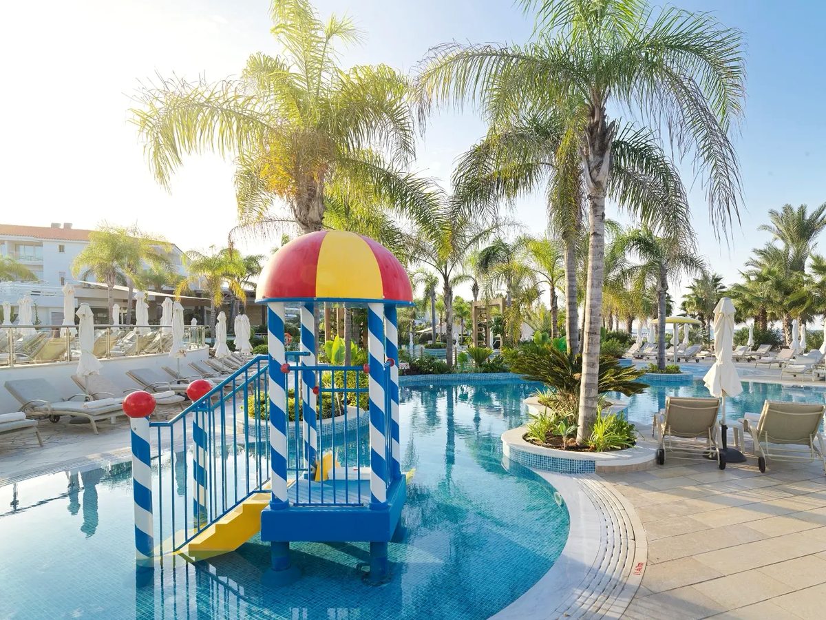 Shallow children's pool with a blue and white play frame, a small yellow slide and a red and yellow canopy, ringed by palms and sun loungers.
