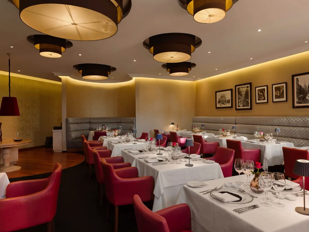 Restaurant with white-linen tables set for dinner, raspberry velvet chairs, banquette seating and drum ceiling lights.
