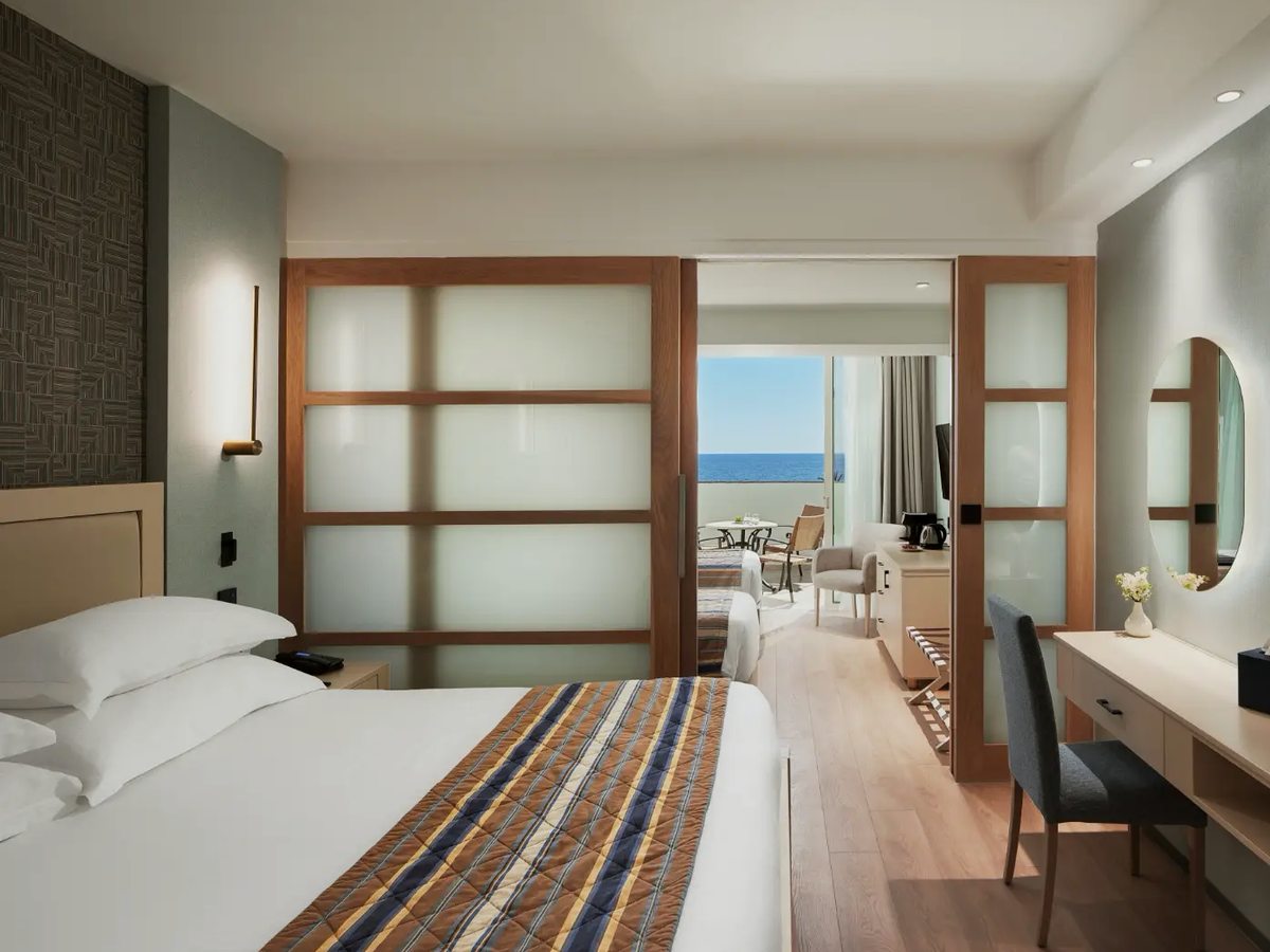 Suite with a double bed in the foreground, a timber-framed frosted sliding partition alongside it and a second sleeping area and balcony with a sea view beyond.