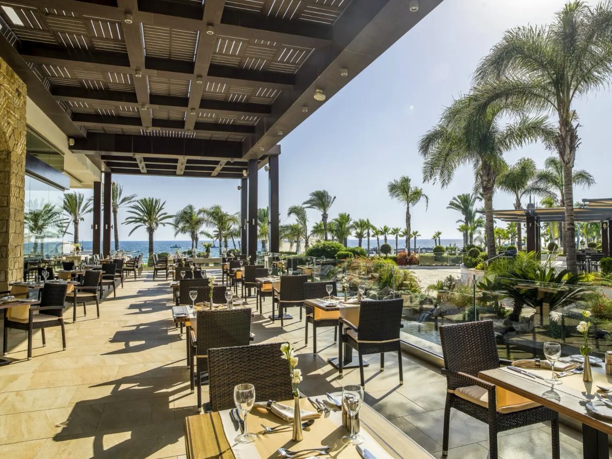 Covered outdoor dining terrace under a slatted pergola, with laid tables and wicker chairs, tall palms beyond and the sea on the horizon.