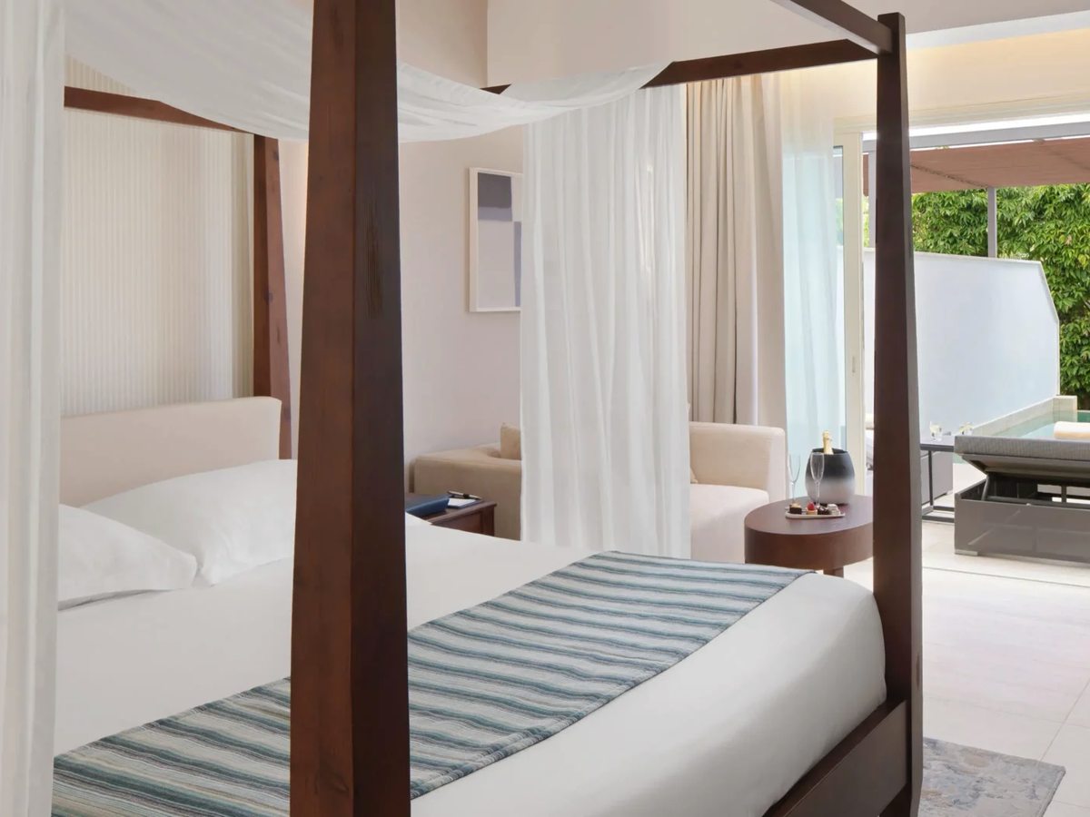 Dark-wood four-poster bed draped with white voile, with a sofa beyond and a private terrace with a plunge pool and sun lounger through the glass doors.