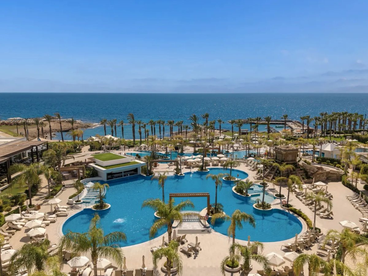 Aerial of the resort's lagoon pools and palm gardens with the open sea and the rocky shoreline beyond.
