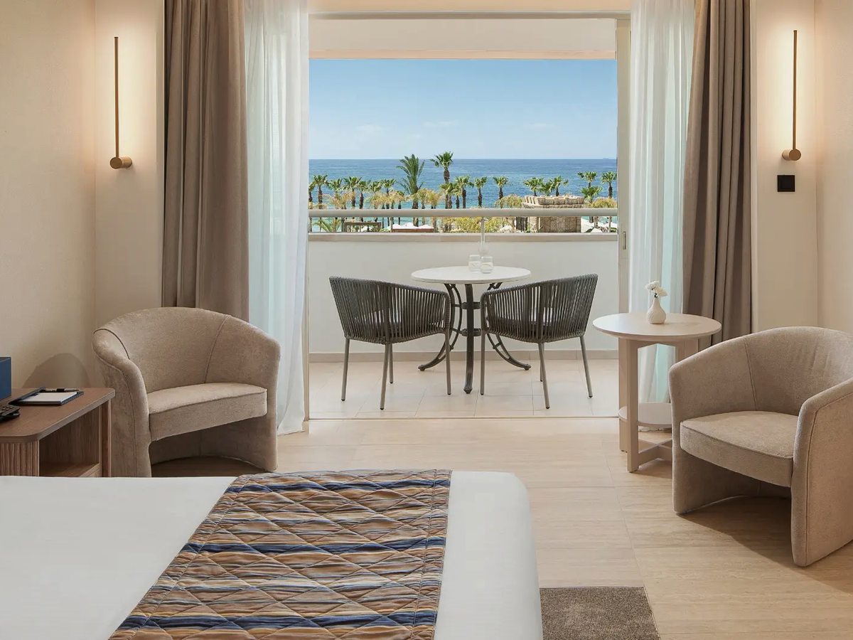 Room with the foot of a bed in front of open balcony doors, two armchairs and a small table, and a furnished balcony looking over palms to the sea.