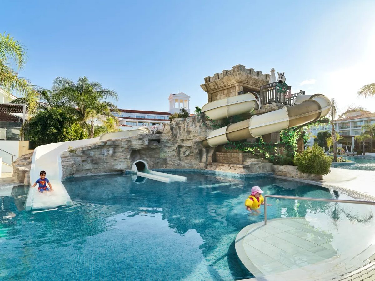 Two enclosed water slides curving down from a stone temple structure into a shallow pool, with a short open slide alongside where a child is coming down and another child standing in the water in a buoyancy vest.