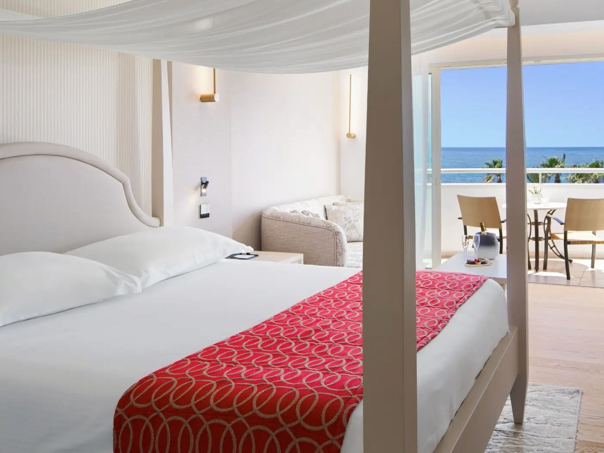Four-poster bed with a white upholstered headboard and a red patterned runner, voile drapes at the posts and a balcony with sea view beyond.
