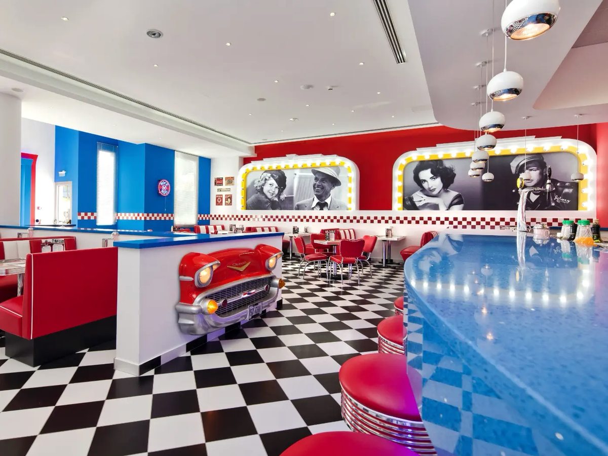 Diner with red leather booths, a black and white chequerboard floor, a counter built from the front of a vintage red car and neon-framed film posters on a blue wall.