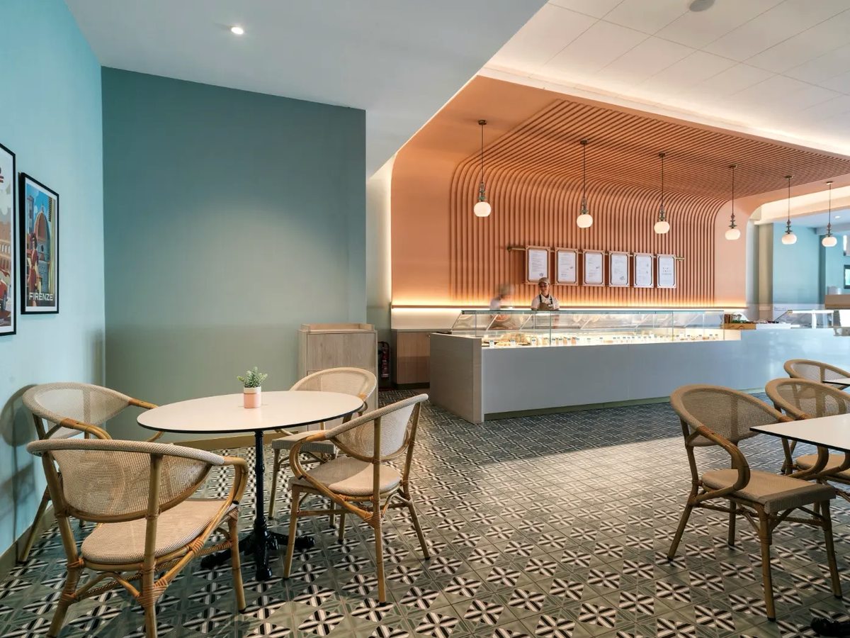 Pale green snack room with round tables and rattan chairs on a patterned tiled floor, facing a lit servery counter with menu boards above.