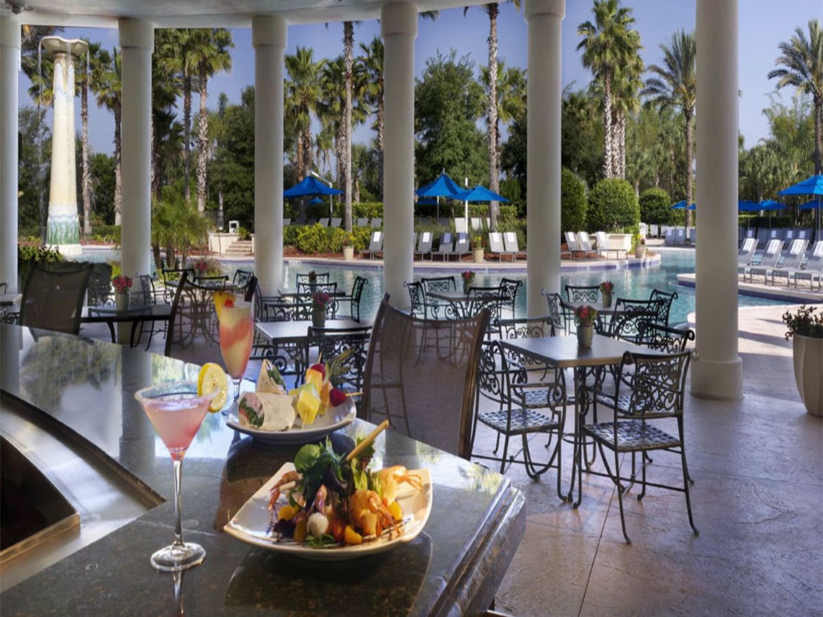A covered outdoor dining terrace with white columns, tables set with cocktails, and a swimming pool with blue parasols and loungers beyond.
