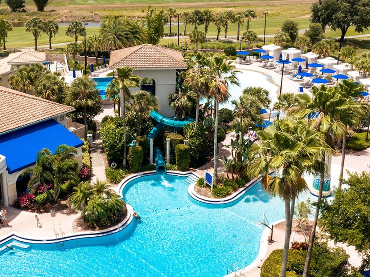 An aerial view of a resort pool complex with a freeform pool, a winding waterslide, loungers, blue umbrellas and tiled-roof poolside pavilions among palms.