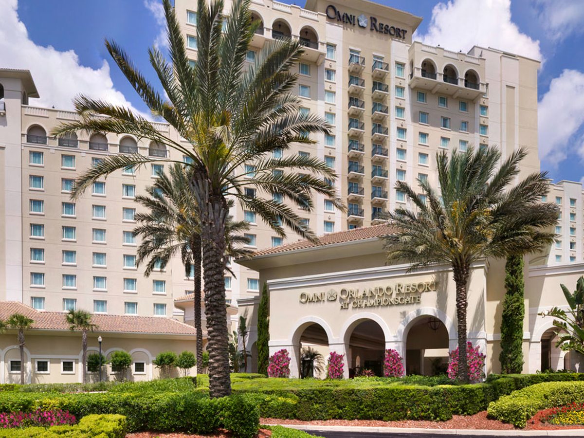 A cream-coloured multi-storey hotel with an arched porte-cochere entrance, framed by tall palm trees and landscaped flowerbeds under a blue sky.