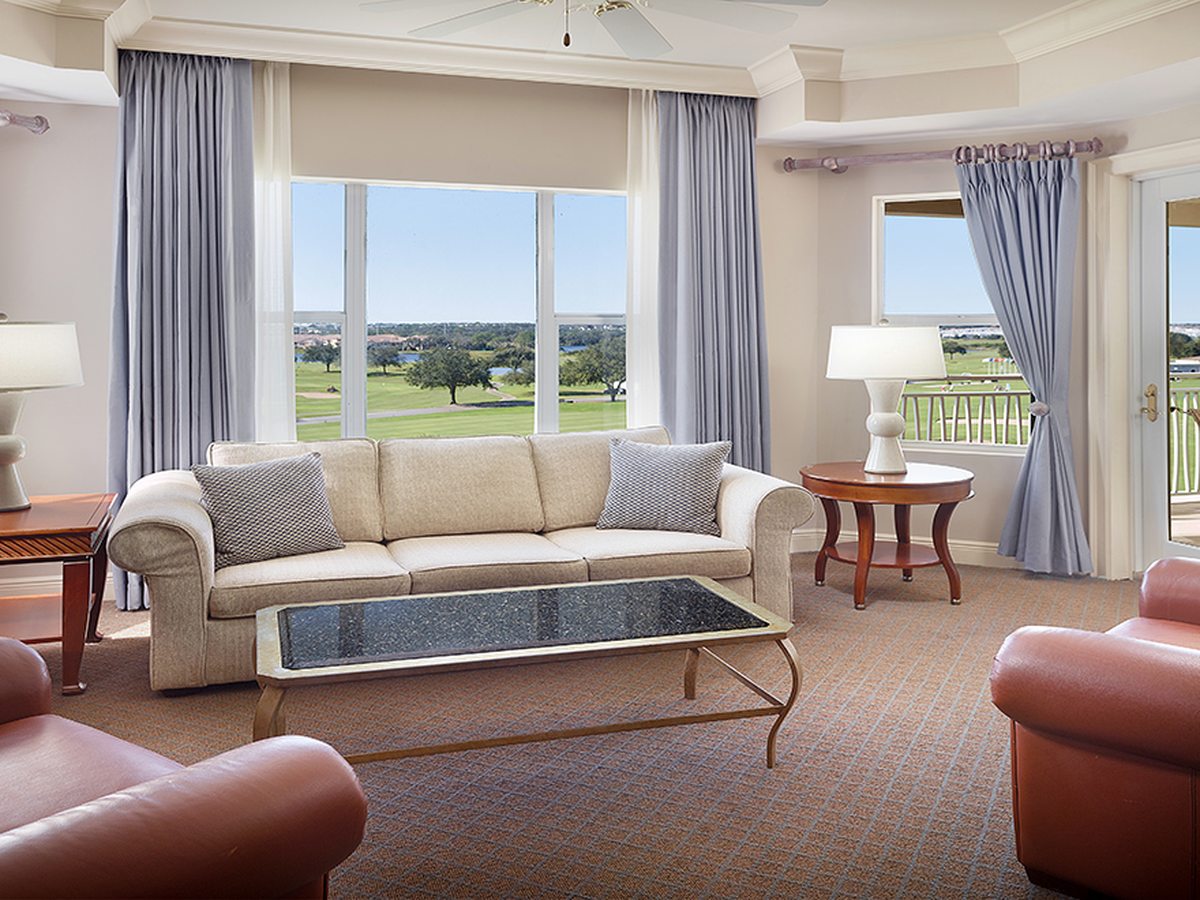 A suite living room with a cream sofa, leather armchairs, a glass coffee table and large windows onto a furnished balcony with a golf-course view.