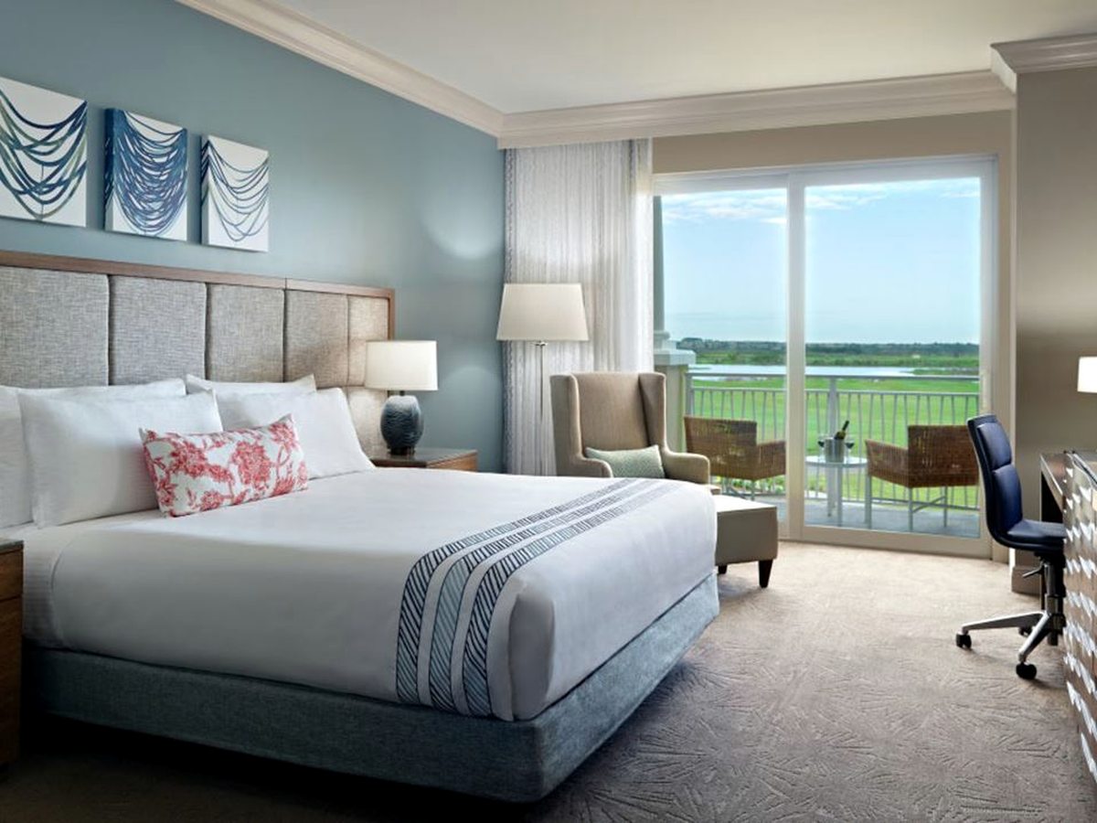 A hotel room with a king bed against a blue accent wall, a work desk, an armchair and sliding doors to a balcony overlooking a golf course.