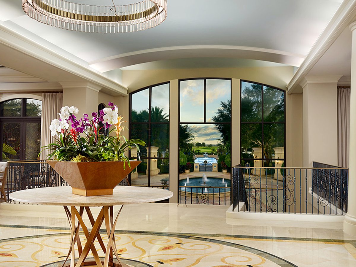 A double-height hotel lobby with cream columns, a crystal chandelier, a round table holding an orchid arrangement and a patterned stone floor.