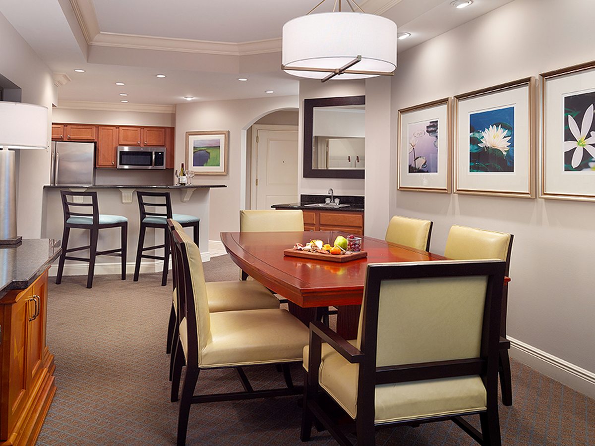 A suite dining area with a rectangular table and upholstered chairs beside a kitchenette with wood cabinets, a microwave and a breakfast bar with stools.