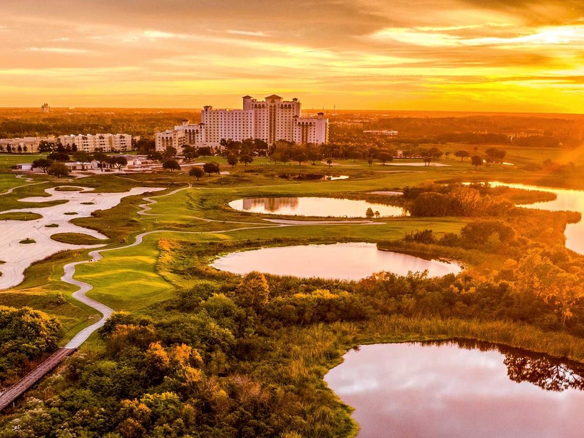 An aerial view at sunset of a large resort spread over golf fairways, bunkers, ponds and wooded grounds, the sky glowing orange.