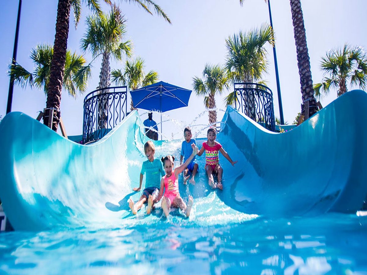 Children riding down a wide open blue waterslide into a shallow landing pool, with an adult watching and palm trees behind.