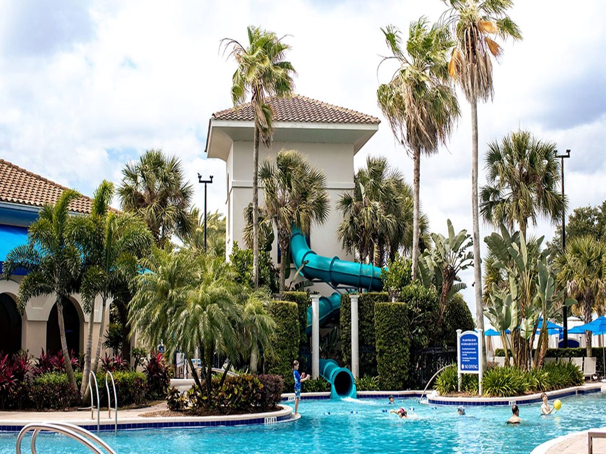 A family pool with a tall enclosed corkscrew tube waterslide, children swimming, pool steps, handrails and palm trees.