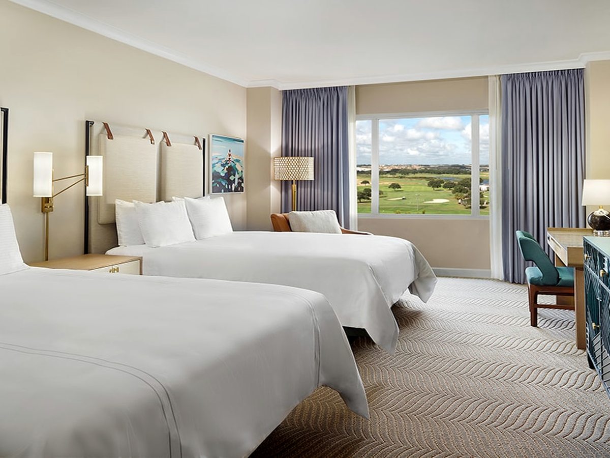 A hotel room with two queen beds, padded headboards, a desk and a large window looking onto a golf course.