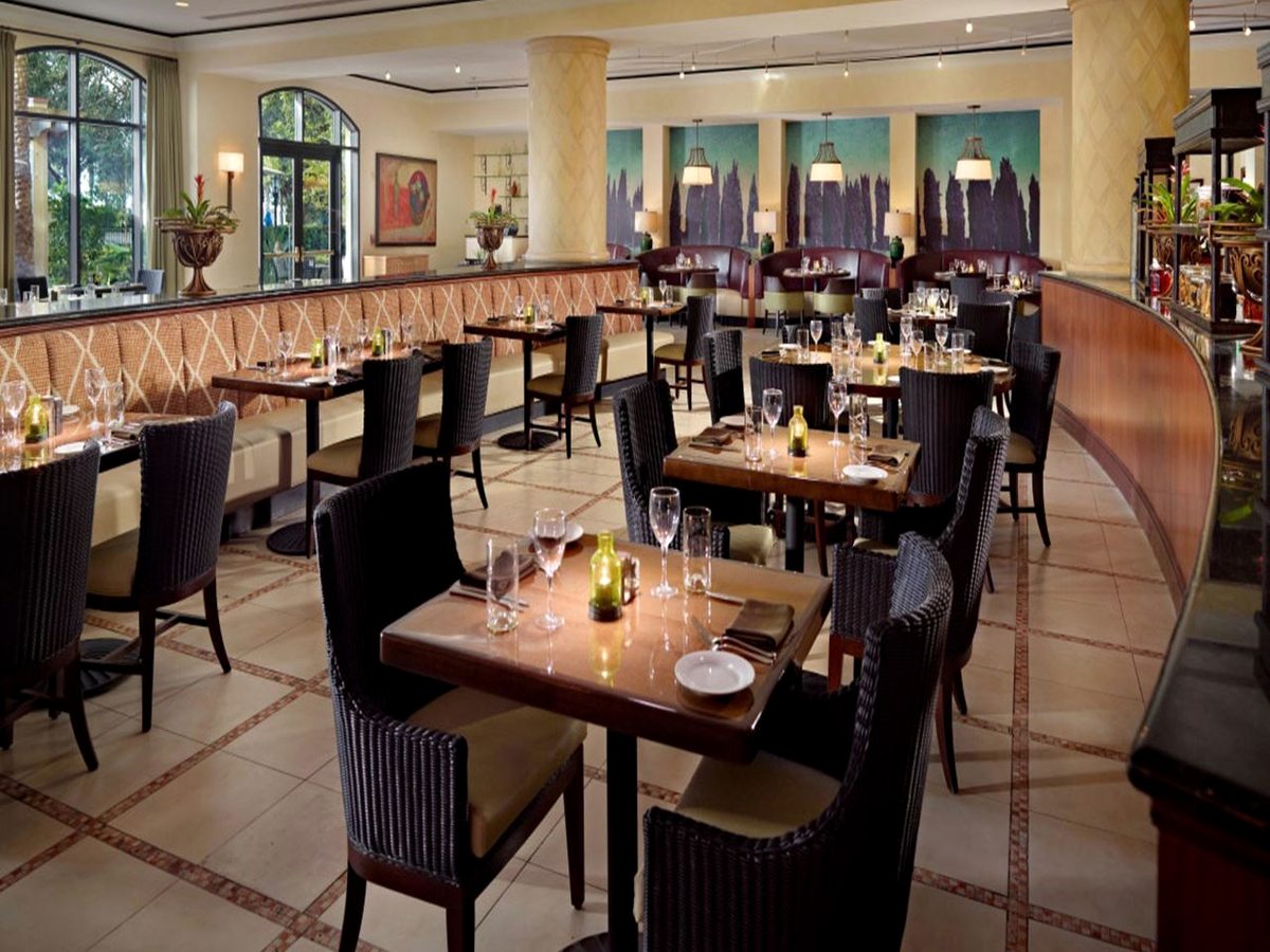 A bright restaurant with set tables, woven chairs, a curved banquette, cream columns, arched windows and a tiled floor.
