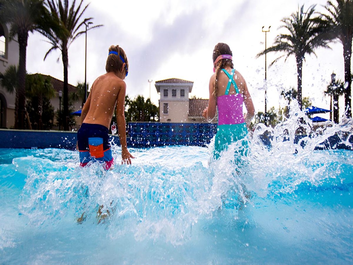 Children splashing in a turquoise wave pool with churning water, palm trees and resort buildings behind.