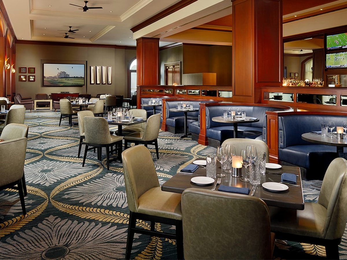 A restaurant with blue upholstered banquettes, cushioned chairs, dark wood partitions and framed golf artwork.